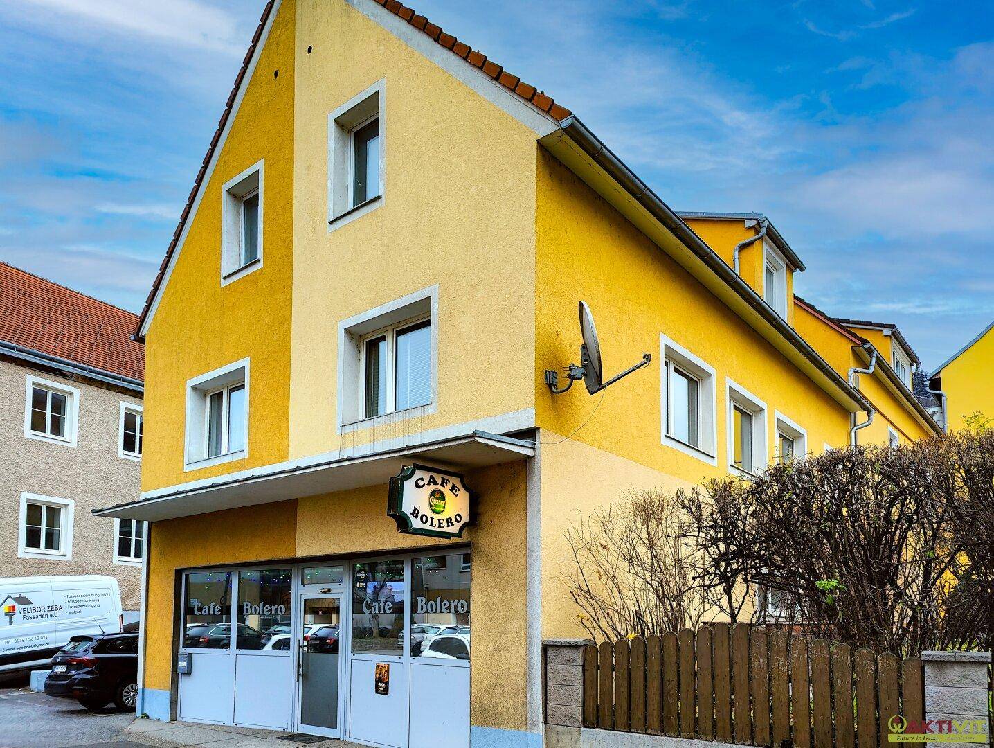 Gelbe Fassade eines Gebäudes mit einem Café im Erdgeschoss und Wohnungen darüber, umgeben von einer Hecke.
