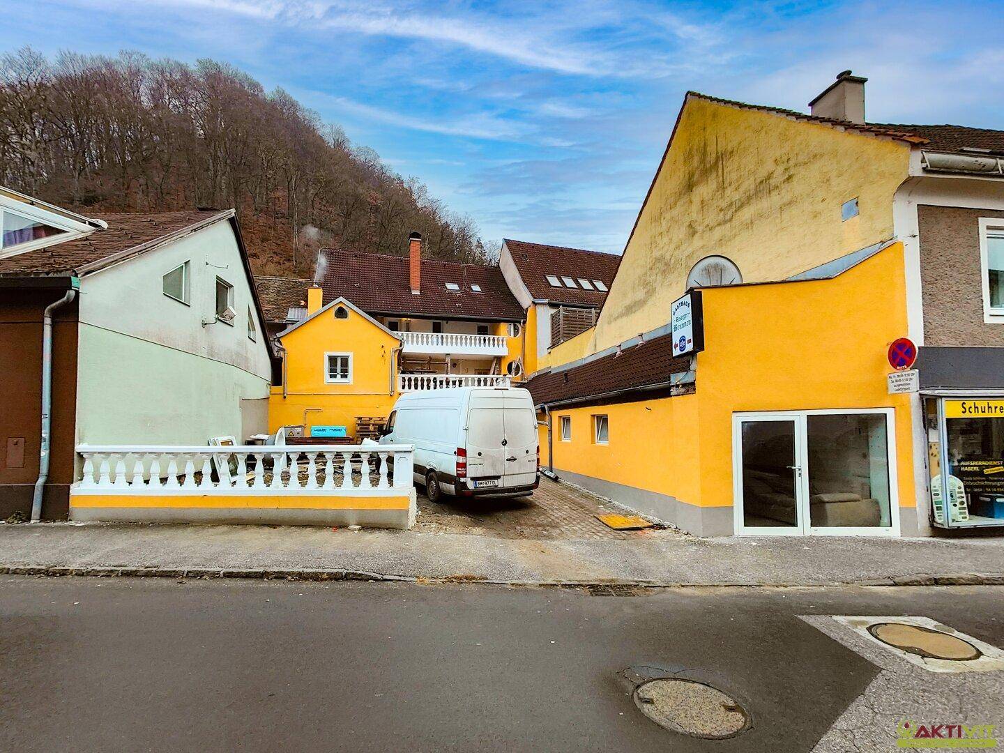 Seitenansicht des gelben Gebäudes mit Parkplatz und umliegenden Häusern vor einem bewaldeten Hügel.