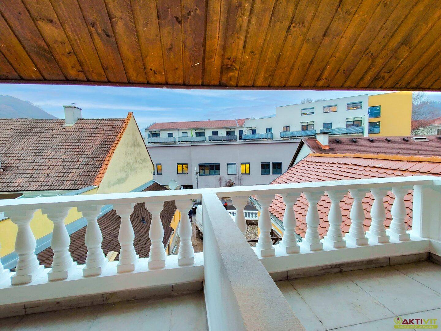 Balkon mit weißem Balustradengeländer und Blick auf die Dächer der Nachbargebäude und einen grünen Hügel.