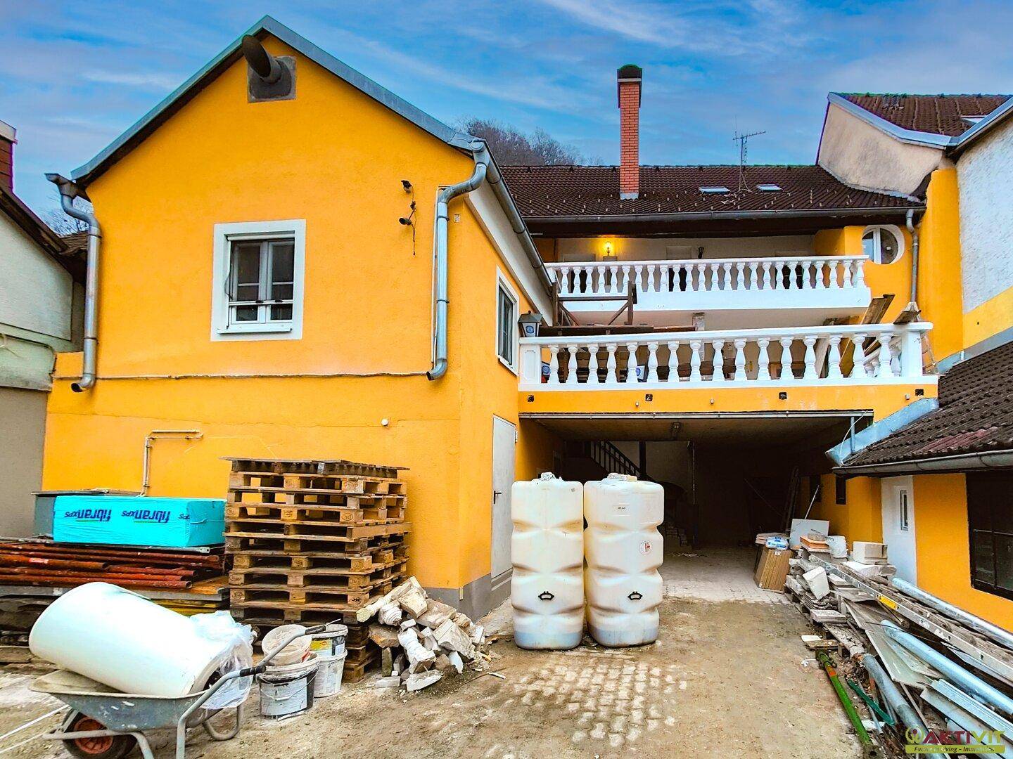 Rückseite des gelben Gebäudes mit einem oberen Balkon, Baustoffen und einem überdachten Durchgang.