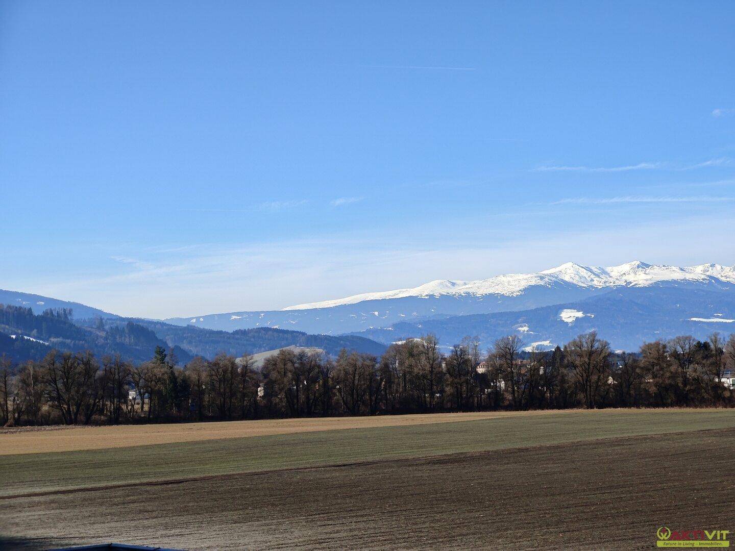 Breiter Ausblick auf eine weite Landschaft mit Feldern, Wäldern und schneebedeckten Bergen am Horizont.