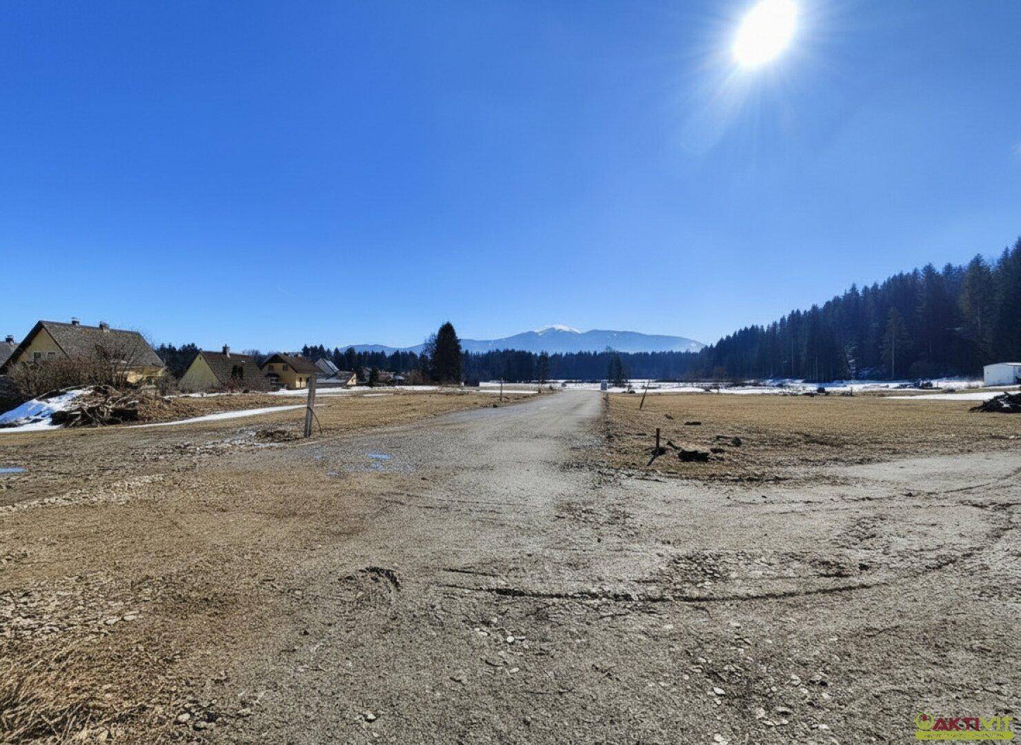Unbefestigter Weg durch weite Felder mit Häusern im Hintergrund und Bergblick.