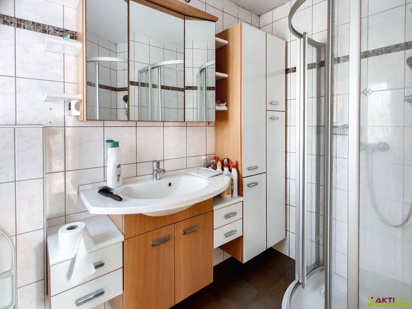 Badezimmer mit Waschtisch, Spiegelschrank, Duschkabine und weißen Fliesen.