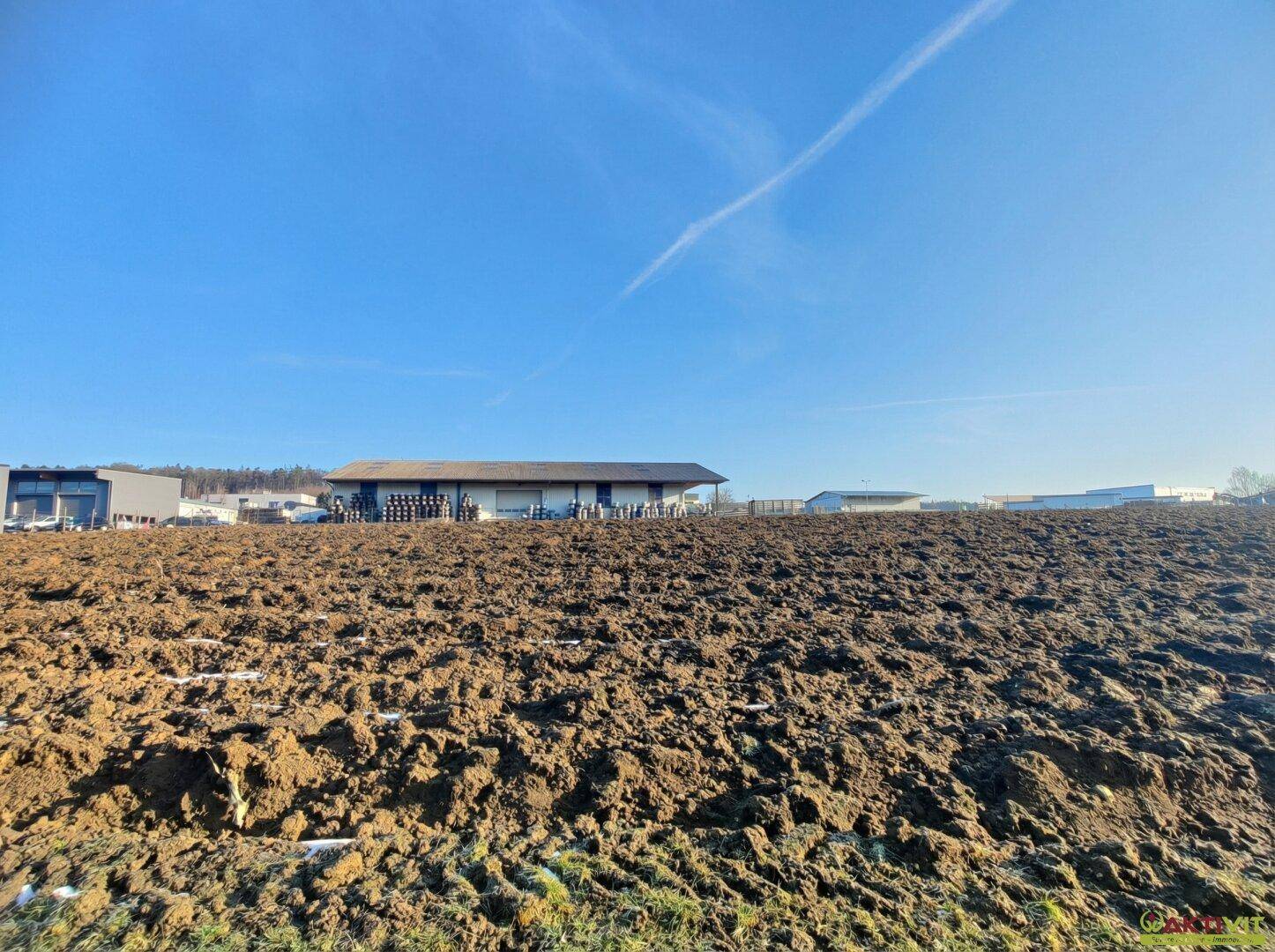 Gepflügtes Feld im Vordergrund mit modernen Industriegebäuden und einem weiten blauen Himmel im Hintergrund.