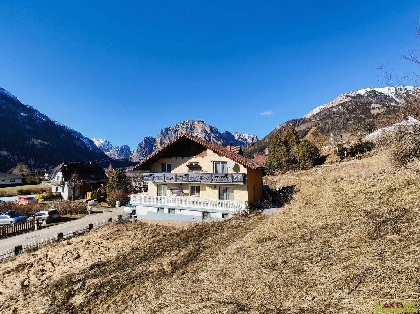 Wohnhaus in alpiner Umgebung mit Bergblick und sonnigem Wetter, umgeben von trockener Vegetation.