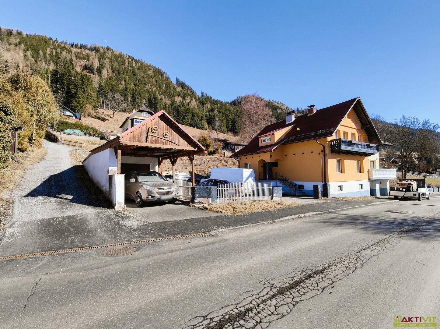 Einfamilienhaus mit Carport und Zufahrt, umgeben von alpiner Landschaft und Bergen.