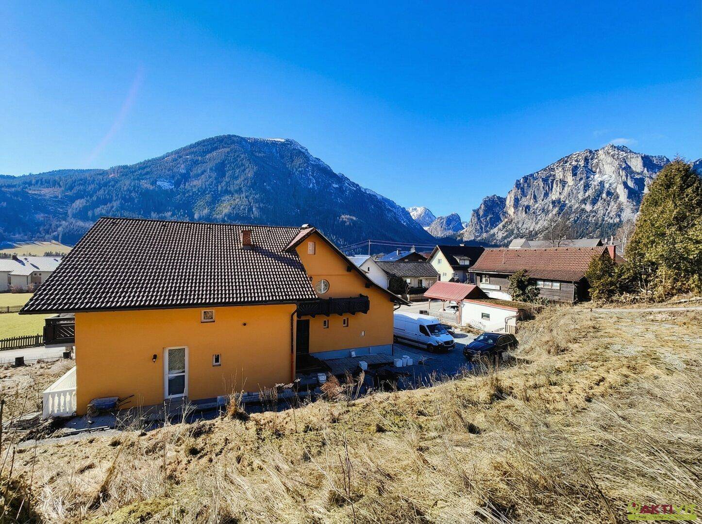 Seitenansicht des Hauses mit Bergblick und umliegender Natur unter blauem Himmel.