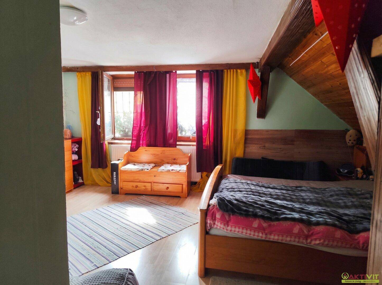Gemütliches Schlafzimmer im Dachgeschoss mit Holzbalken, Holzbett und bunten Vorhängen.