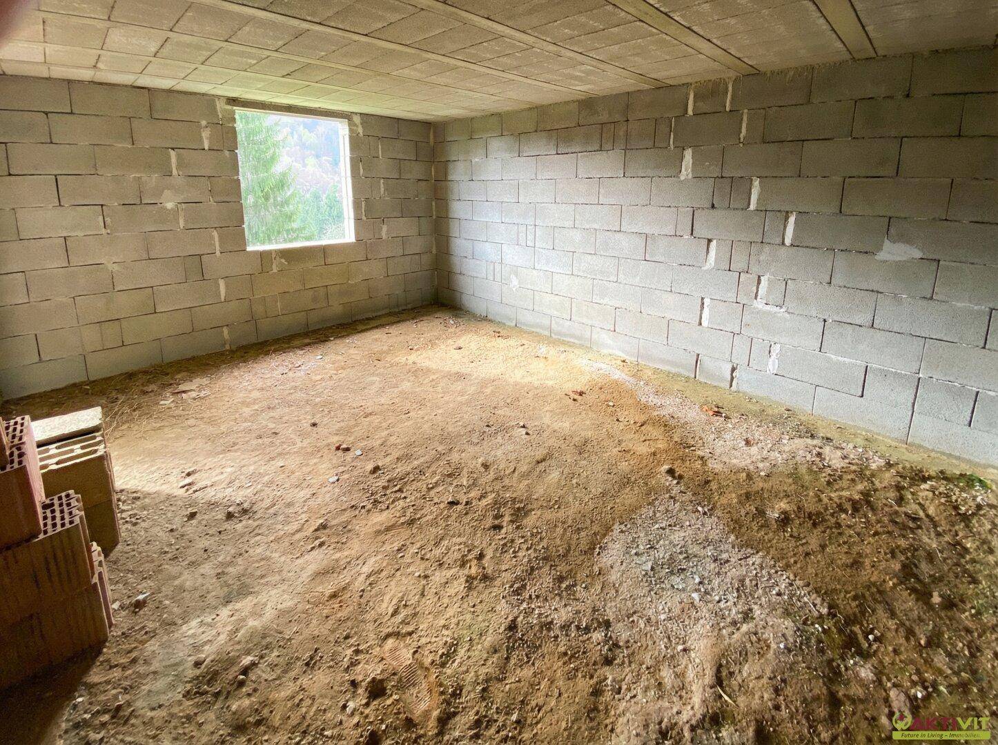 Rohbau-Innenraum mit Betonblockwänden, Erdboden und einem kleinen Fenster mit Blick ins Grüne.