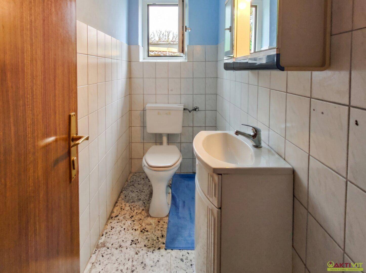 Kleines WC mit Fenster, Waschbecken und weiß gefliesten Wänden.