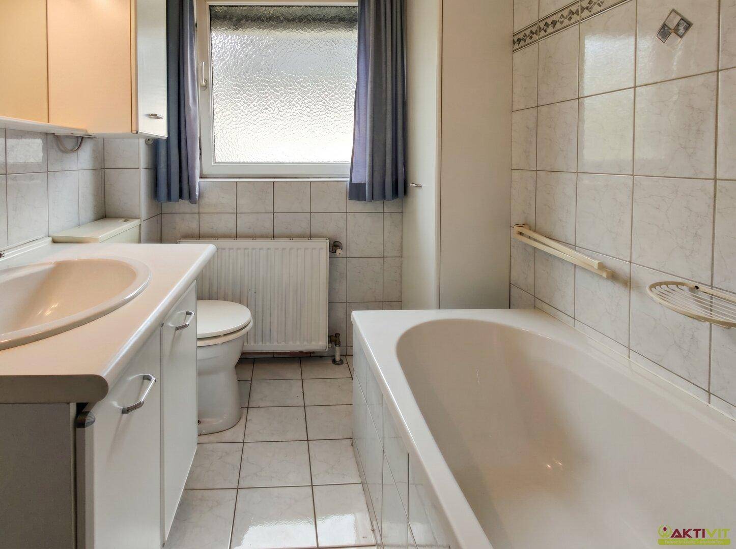Badezimmer mit Badewanne, Waschbecken, WC und Fenster, komplett gefliest.