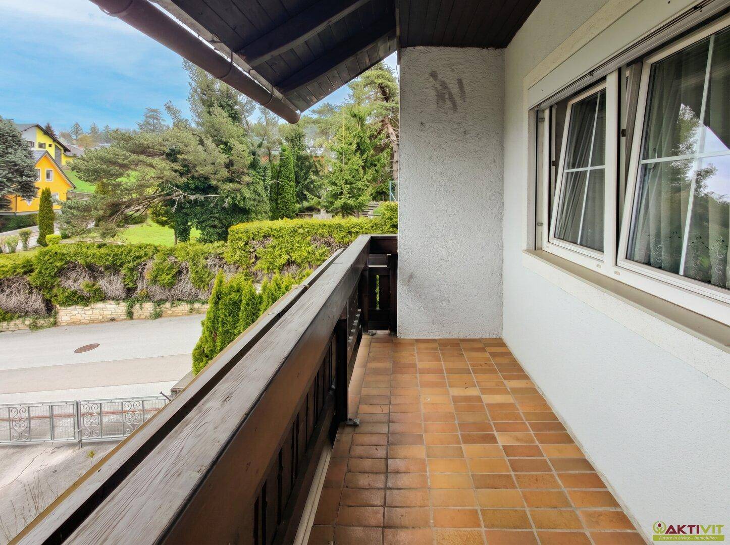 Langer Balkon mit Terrakotta-Fliesen und Holzgeländer, umgeben von Bäumen.