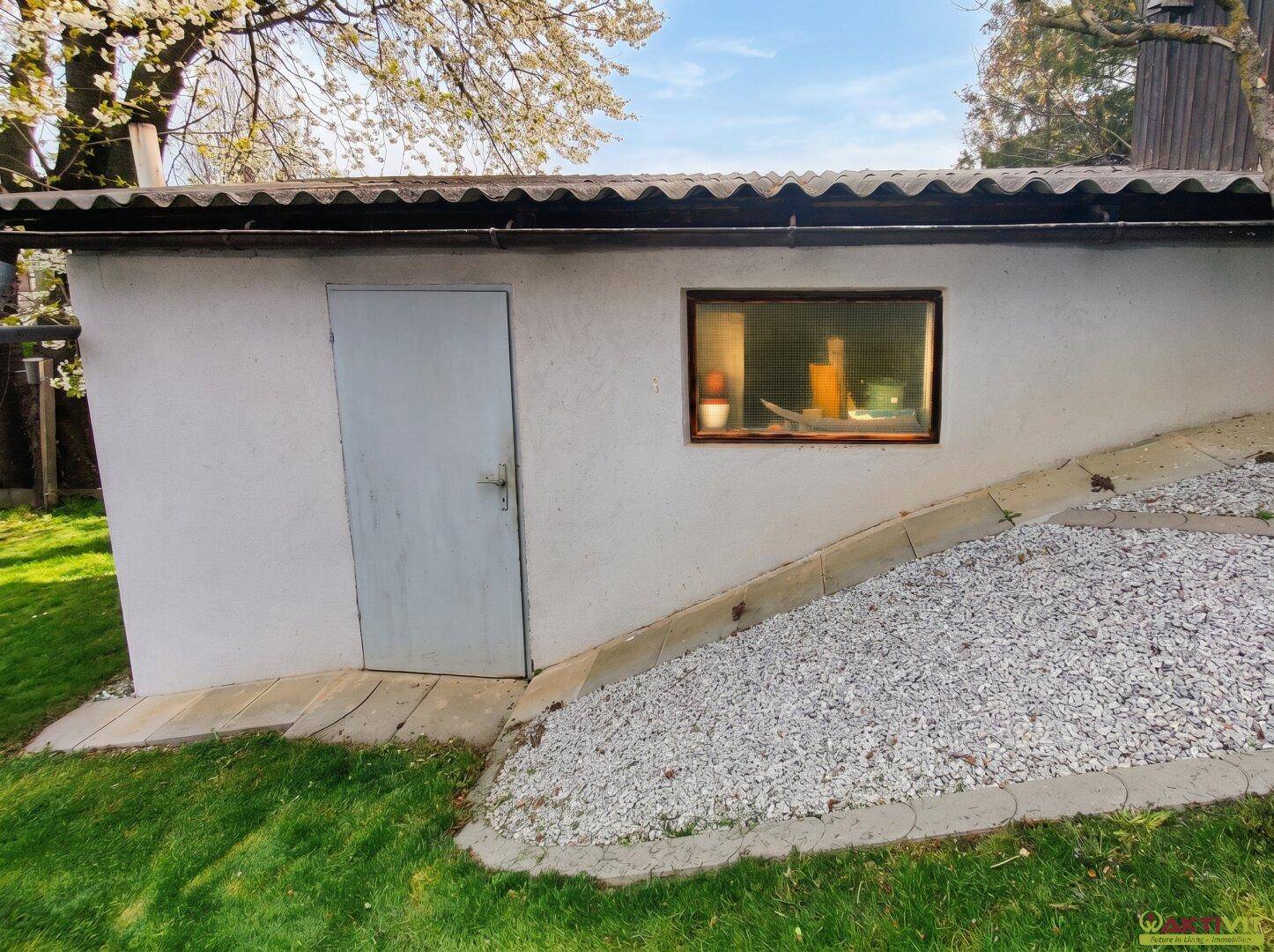 Kleiner Gartenschuppen mit Fenster und Tür, umgeben von Rasen und Kies.