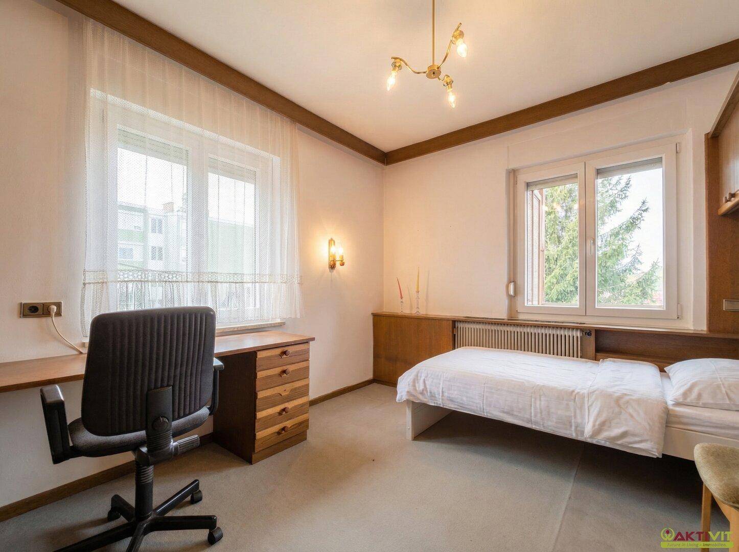 Helles Schlafzimmer mit Einzelbett, Schreibtisch und zwei Fenstern.