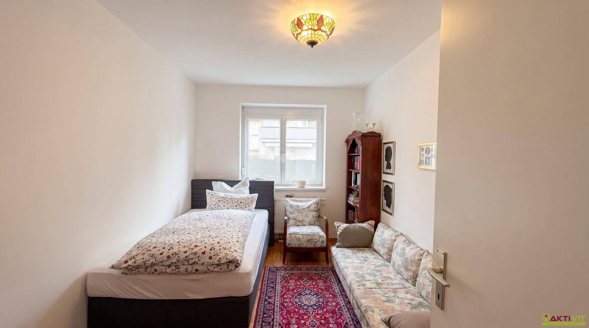 Helles Schlafzimmer mit Einzelbett, floralem Sofa und Fenster zum Außenbereich.