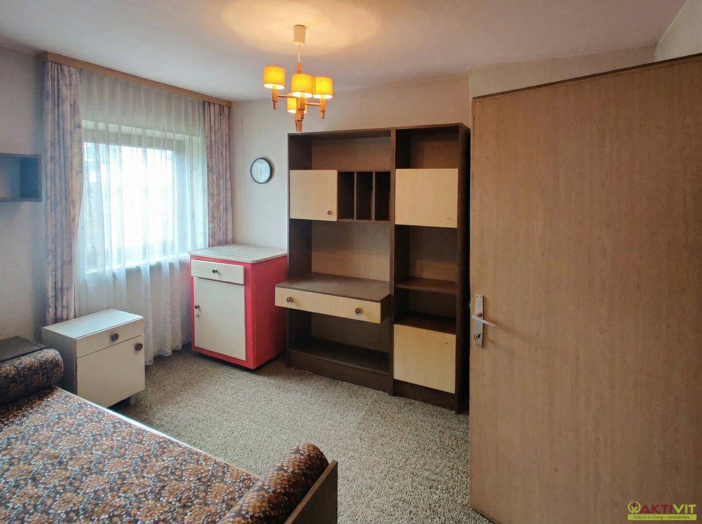 Schlafzimmer mit Schreibtisch, Schrank und Fenster mit Vorhängen.