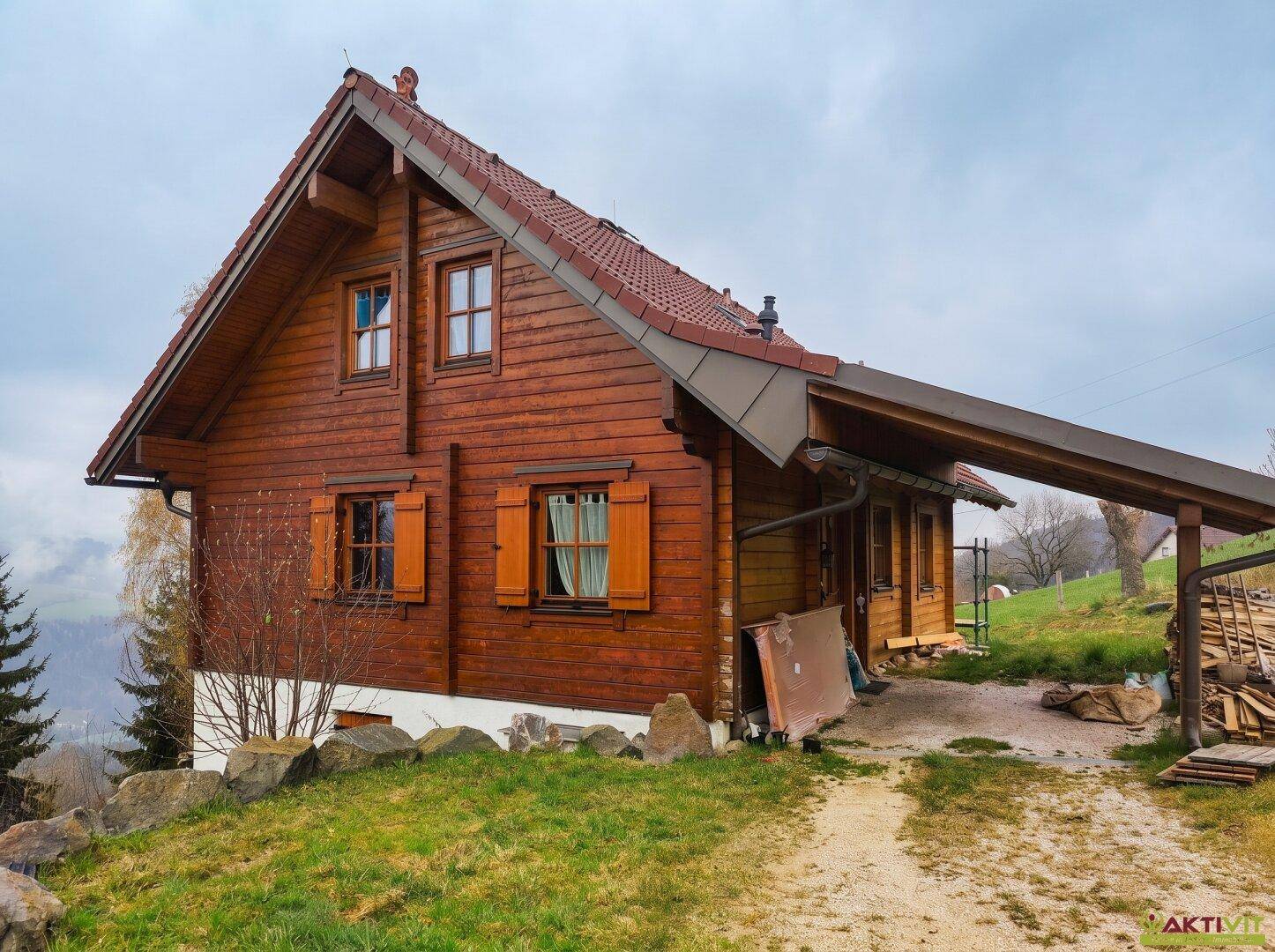 Rustikales Holzhaus mit Satteldach und Carport in ländlicher Umgebung an einem bewölkten Tag.