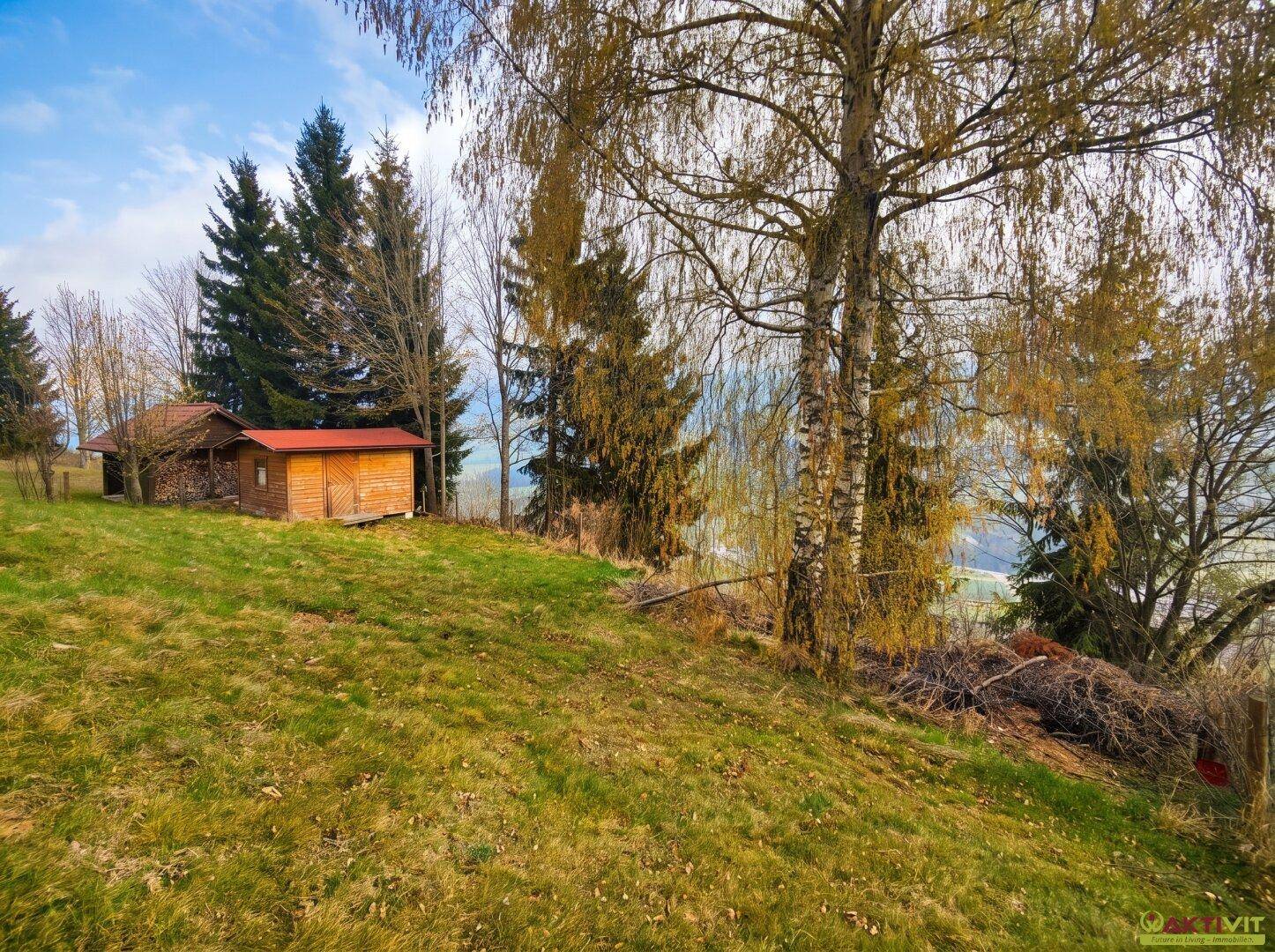 Weitläufige grüne Hügellandschaft mit Bäumen und einem kleinen Schuppen im Hintergrund.