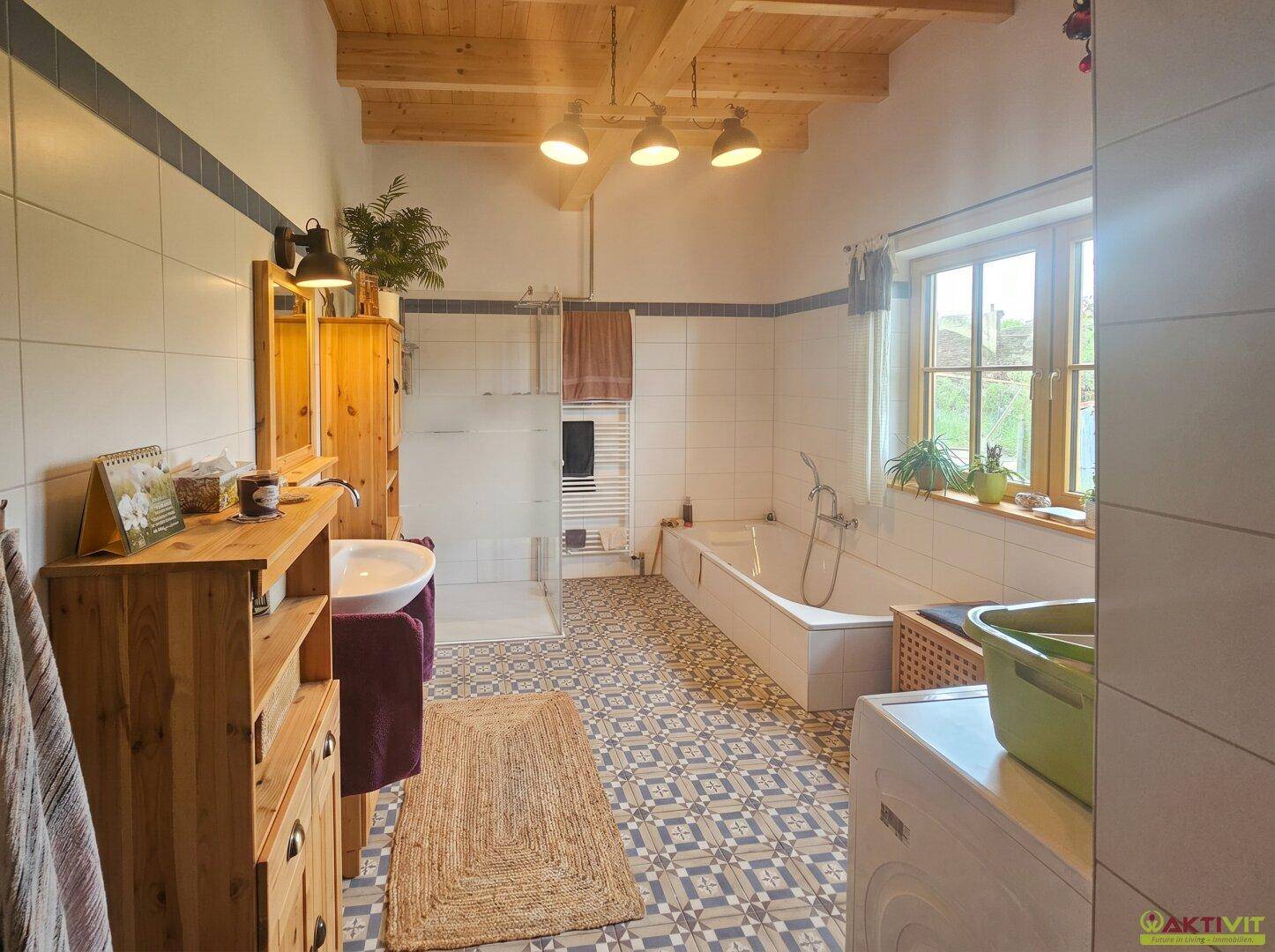 Geräumiges Badezimmer mit Badewanne, Dusche, Fenster und gemusterten Bodenfliesen in hellem Design.