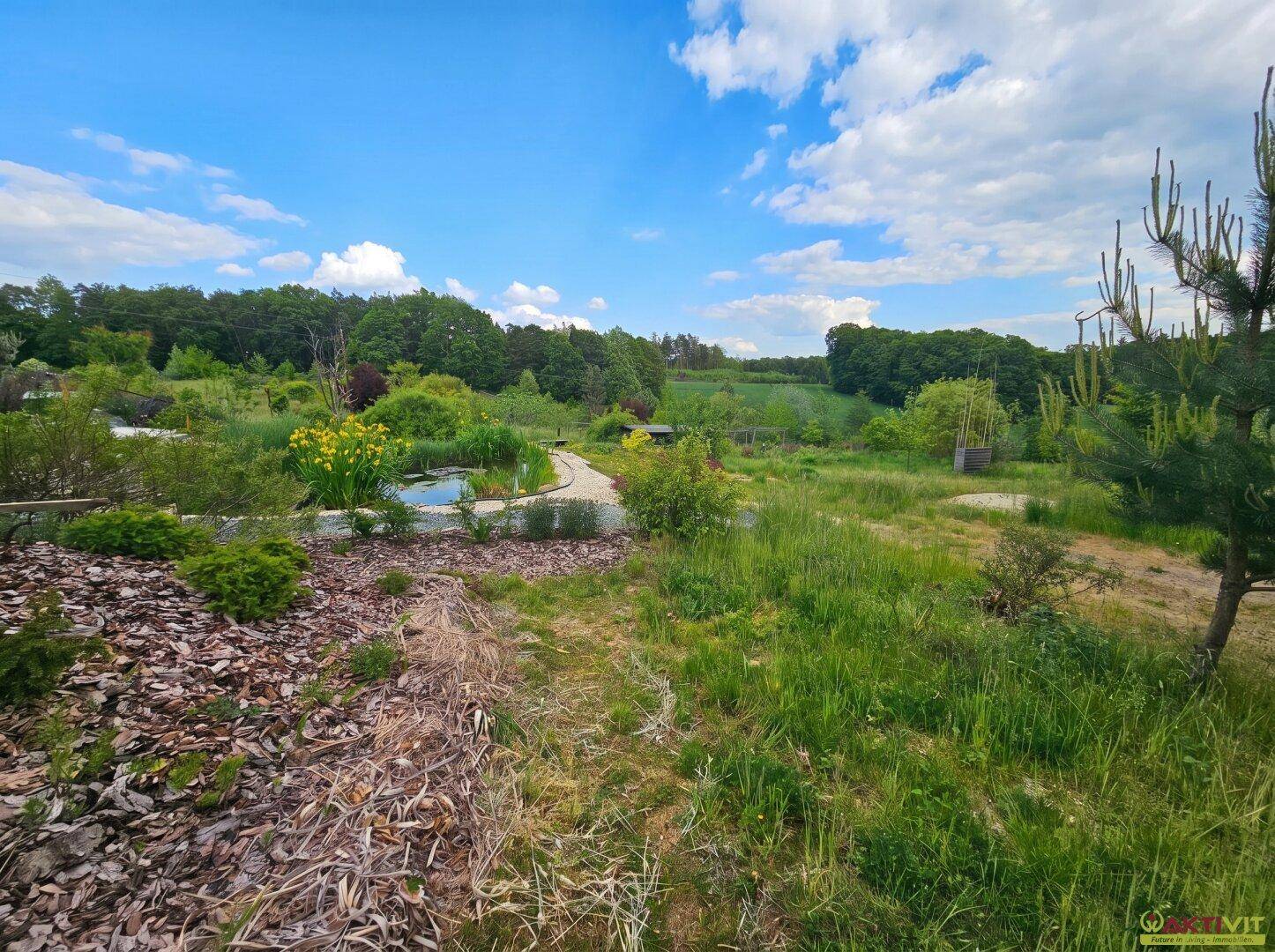 Weitläufiger Gartenbereich mit Wiesen, Wegen und Blick auf den Teich und die umliegende Natur.