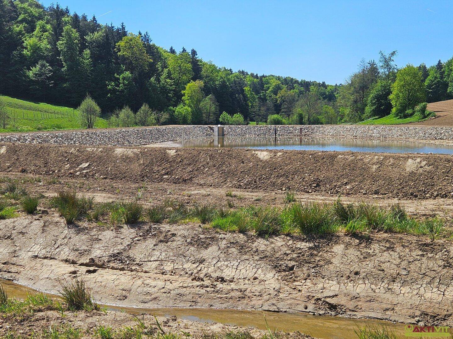 Panoramablick auf einen Wasserspeicher mit Erdarbeiten und spärlicher Vegetation vor einem Waldhintergrund.