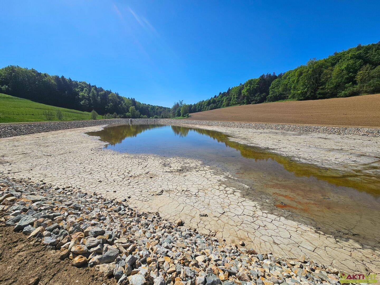 Breite Ansicht eines Teiches mit rissigem Boden im Vordergrund und bewaldeten Hügeln im Hintergrund unter blauem Himmel.