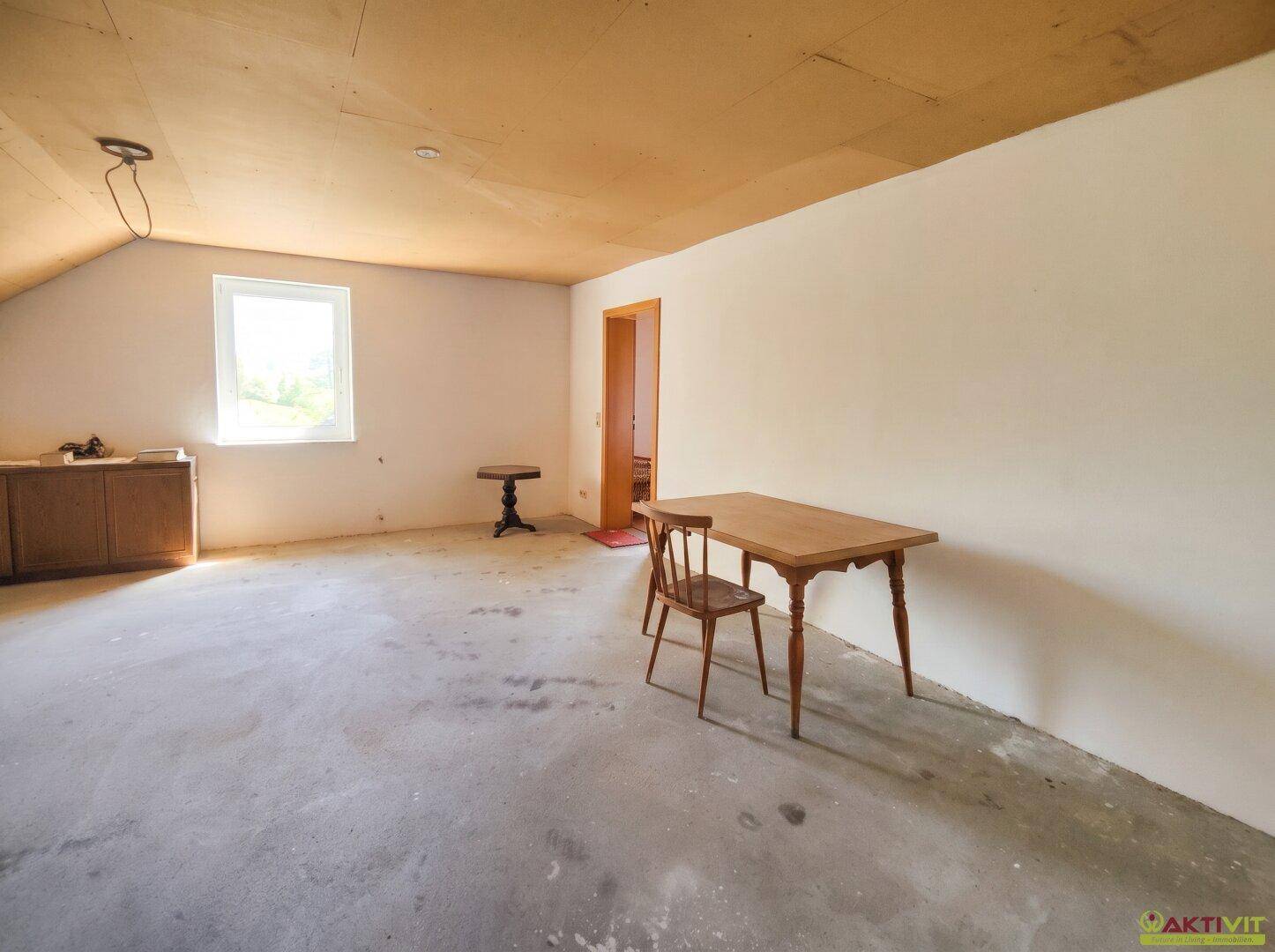 Heller Dachraum mit schräger Decke, Betonboden und einem Fenster, das Tageslicht hereinlässt.