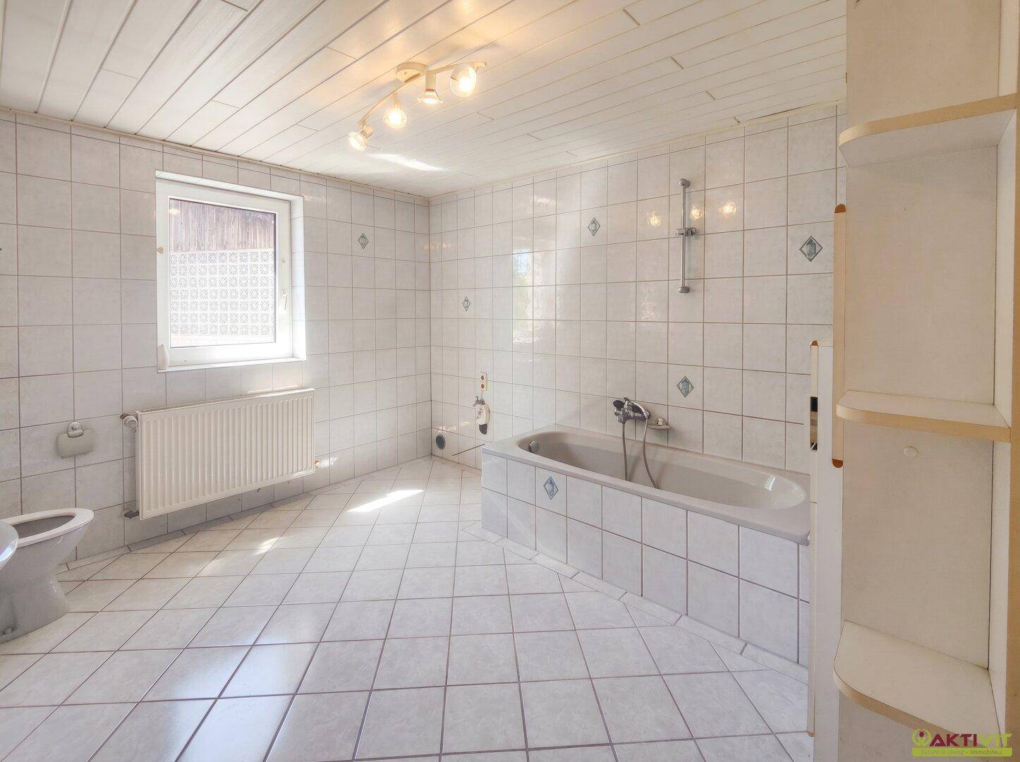 Geräumiges Badezimmer mit weißen Fliesen, Badewanne, Toilette und einem Fenster für natürliche Belüftung.