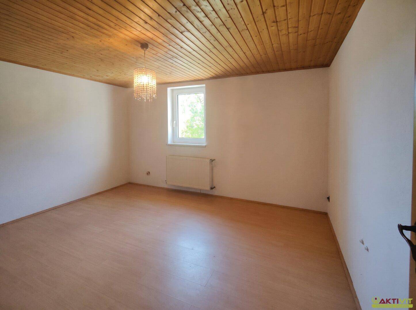 Gemütliches Schlafzimmer mit Holzbalkendecke, Laminatboden und einem Fenster mit Blick ins Grüne.