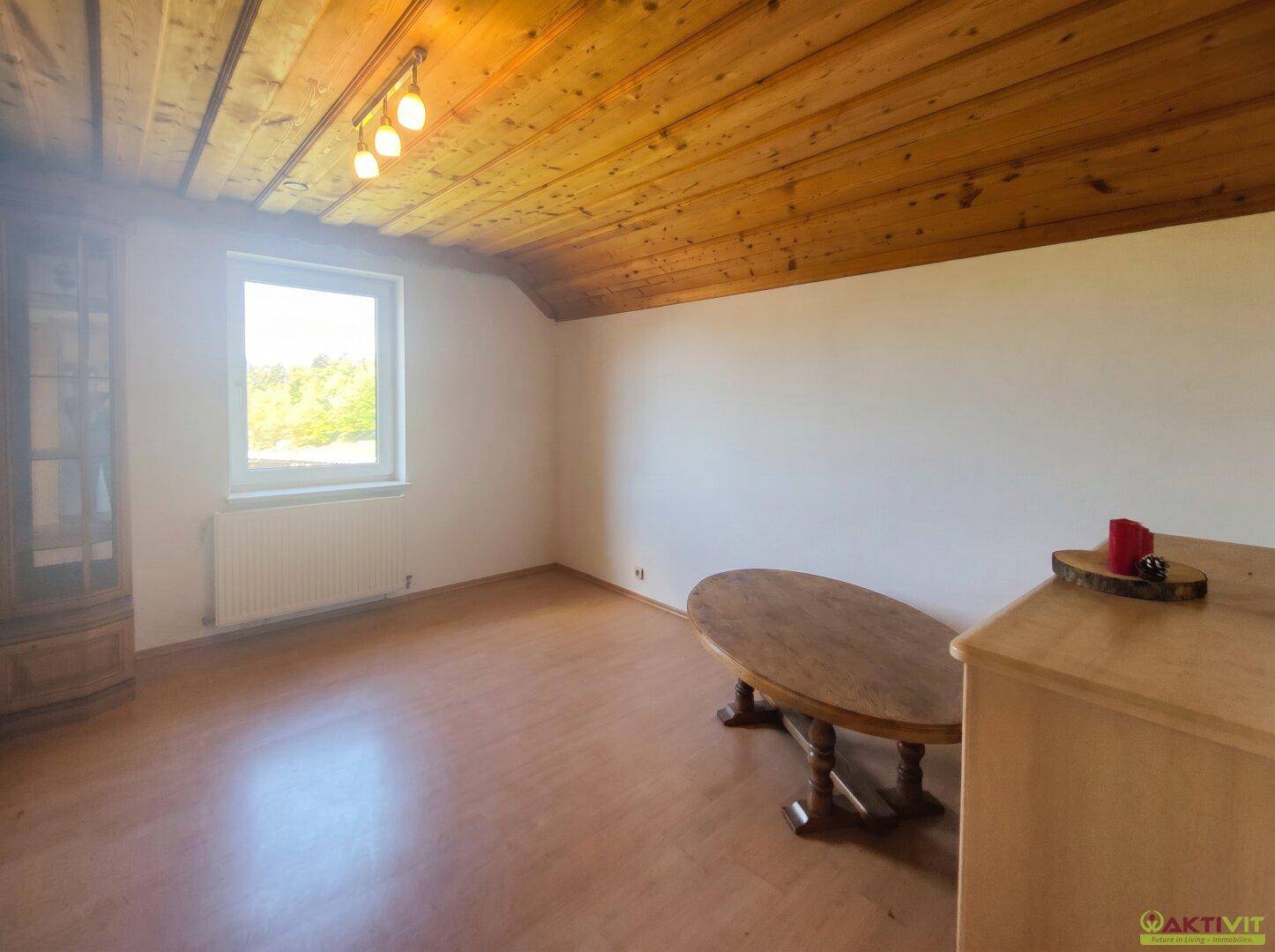 Heller Wohnraum mit Holzbalkendecke, Laminatboden und einem Fenster, das viel Tageslicht hereinlässt.