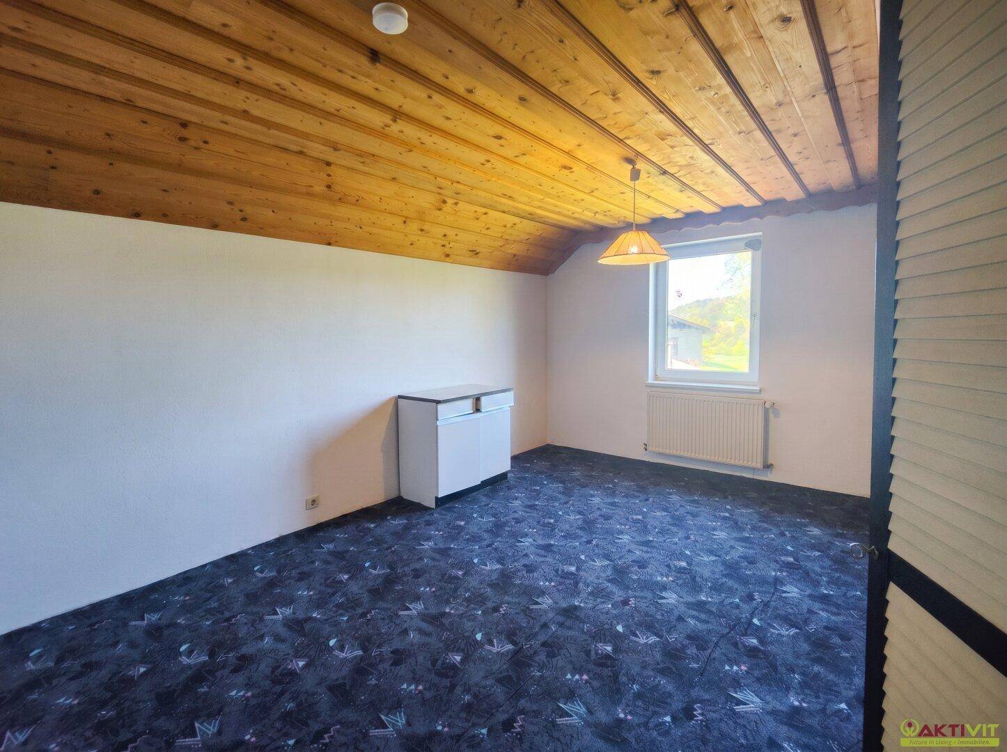 Schlafzimmer im Dachgeschoss mit schräger Holzdecke, Teppichboden und einem Fenster mit Ausblick.