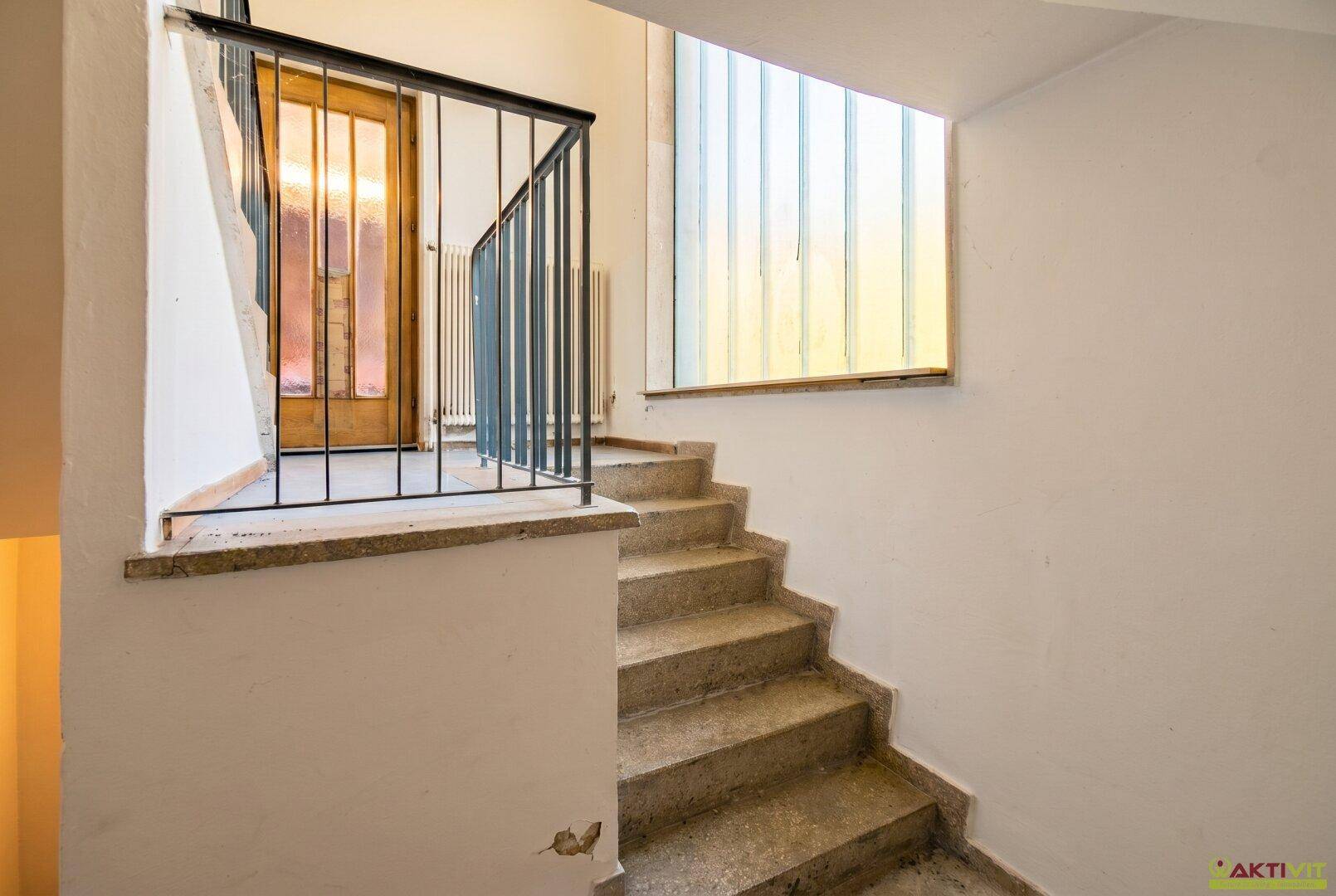 Treppenhaus mit Steinstufen, Metallgeländer und einem Fenster aus Glasbausteinen.
