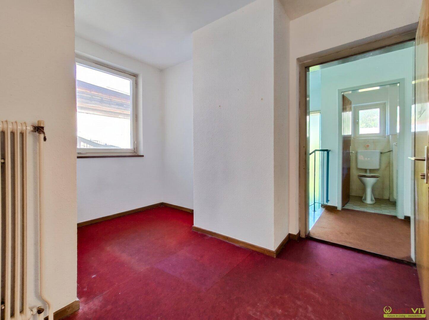 Kleiner Raum mit rotem Teppichboden, Fenster und Heizkörper, angrenzend an ein WC.