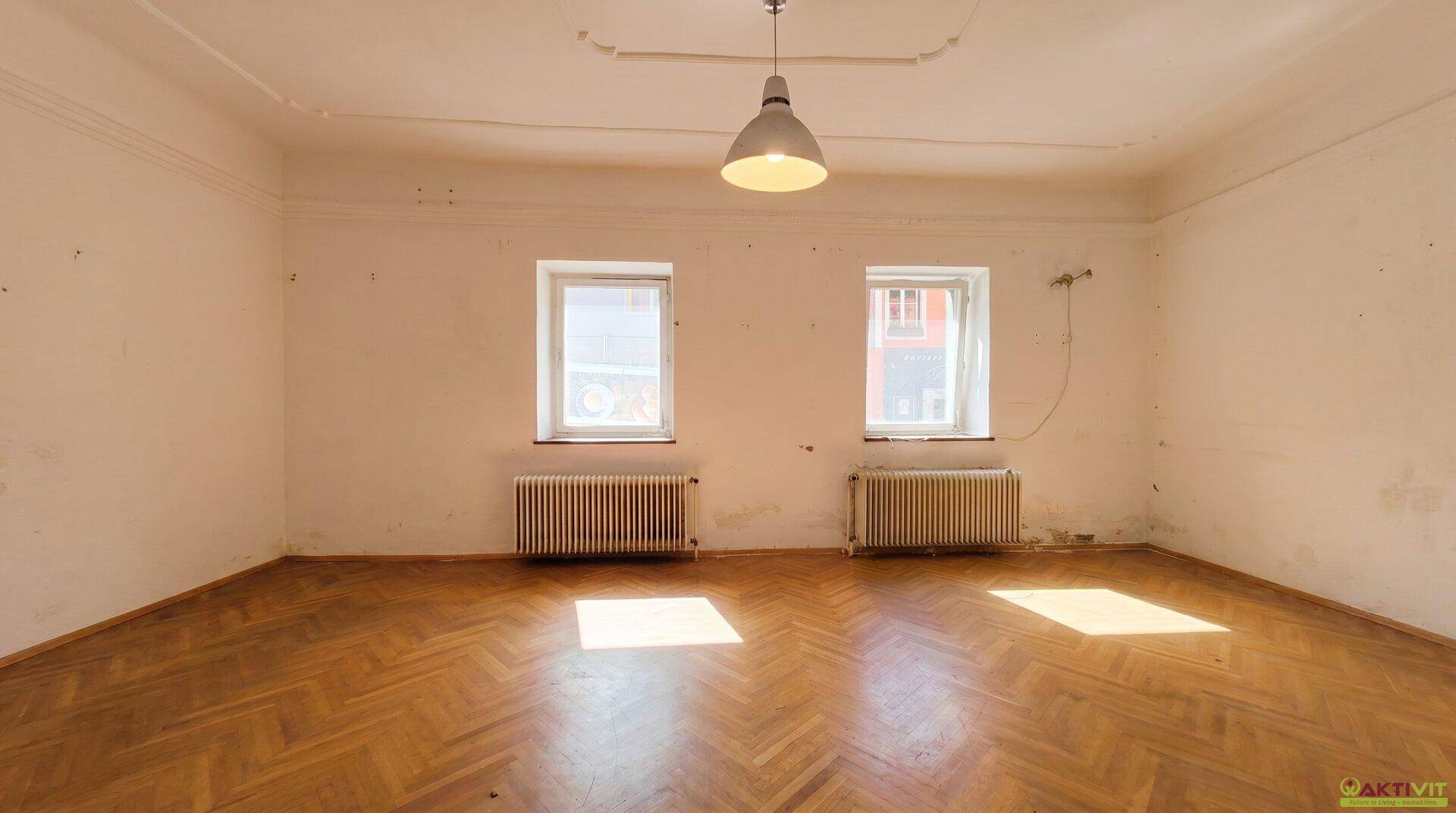 Geräumiges Wohnzimmer mit Fischgrätparkett, zwei Fenstern und Heizkörpern.