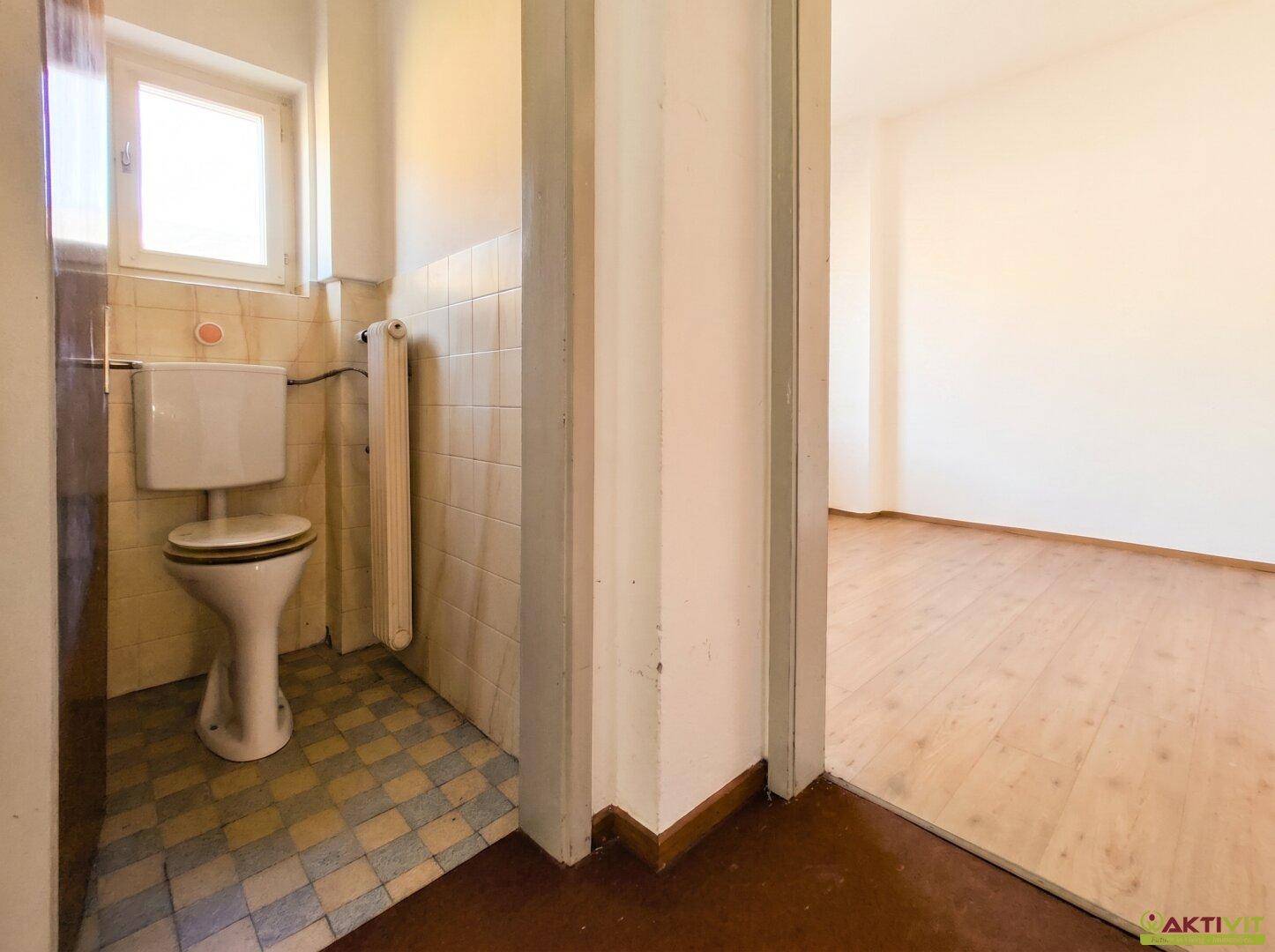 Separates WC mit Toilette, Fenster und gefliestem Boden, angrenzend an einen weiteren Raum.