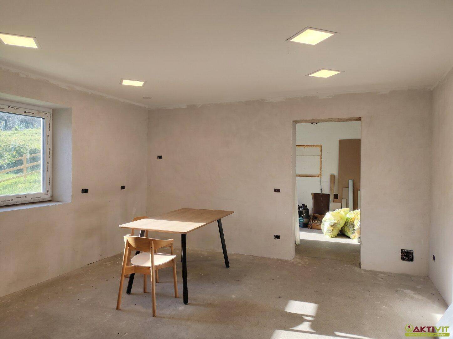 Renovierungsbedürftiger Raum mit neuem Fenster, Holztisch und Einbaustrahlern.