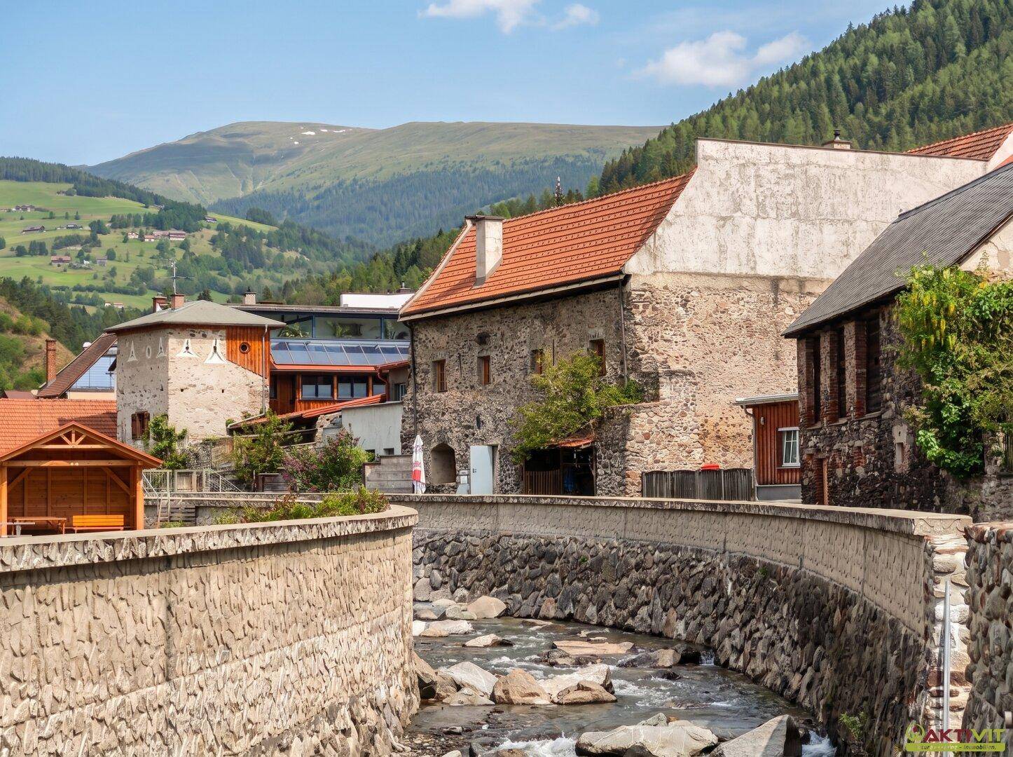 Historische Steingebäude entlang eines Flusses mit grünen Bergen im Hintergrund.