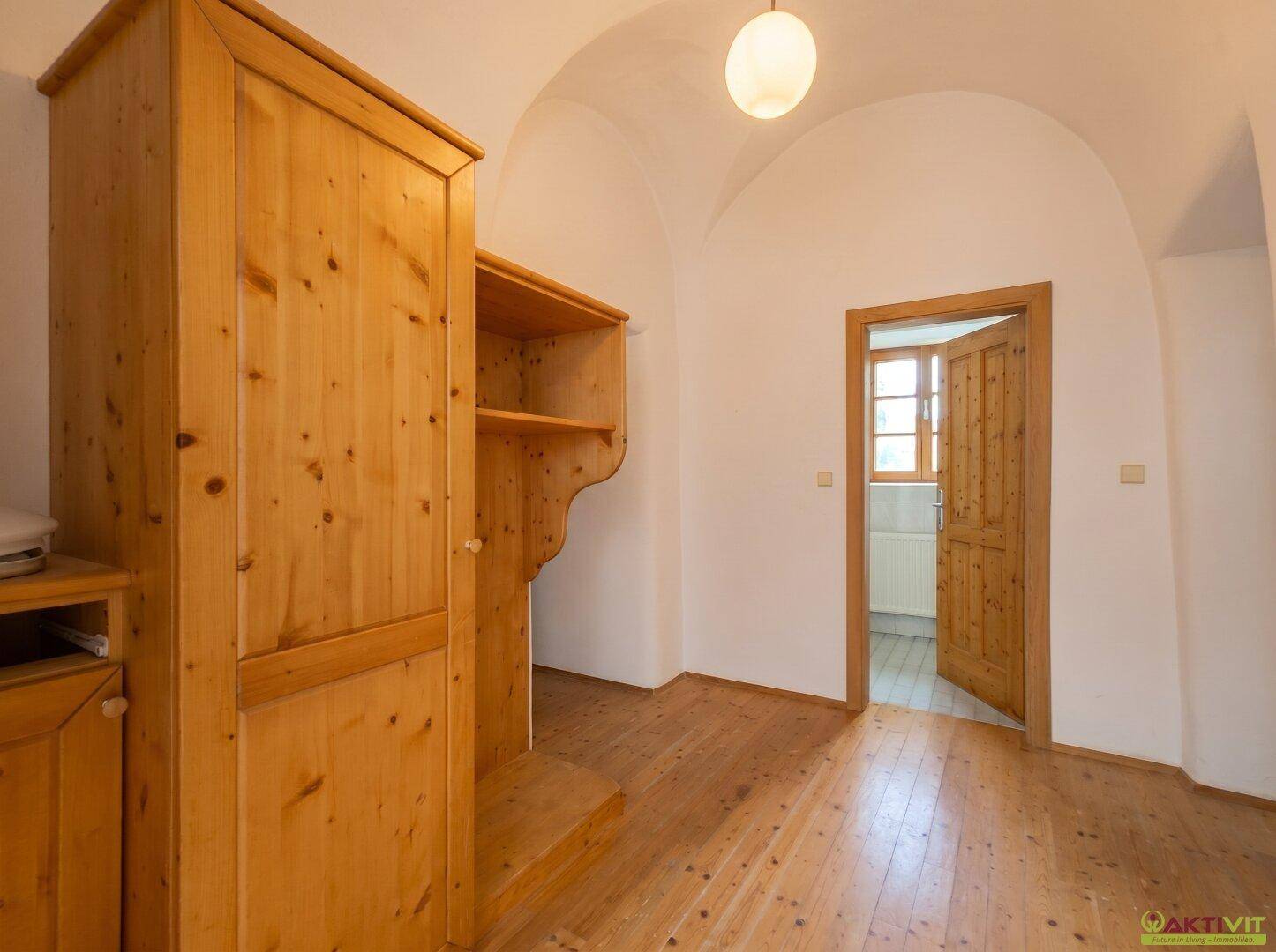 Helles Zimmer mit Holzboden, Holzschrank und offener Tür zu einem angrenzenden Raum.