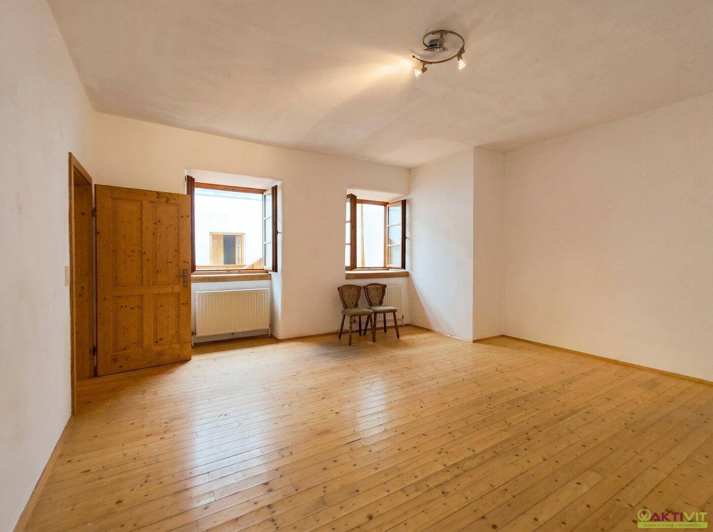 Helles, leeres Wohnzimmer mit Holzboden, zwei Fenstern und einer Holztür.