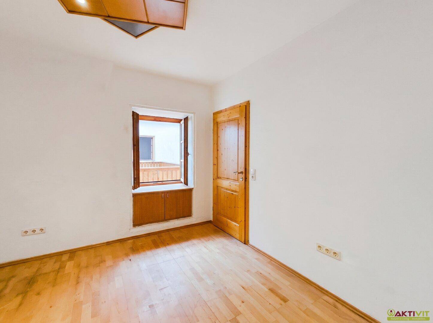 Leeres Zimmer mit Holzboden, Holztür und einem Fenster mit Blick nach draußen.