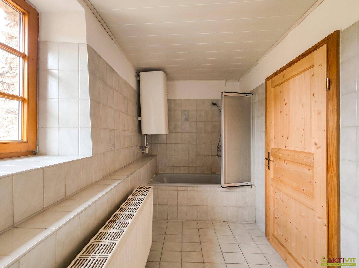Gefliestes Badezimmer mit Badewanne, Duschvorrichtung, Fenster und Heizkörper.