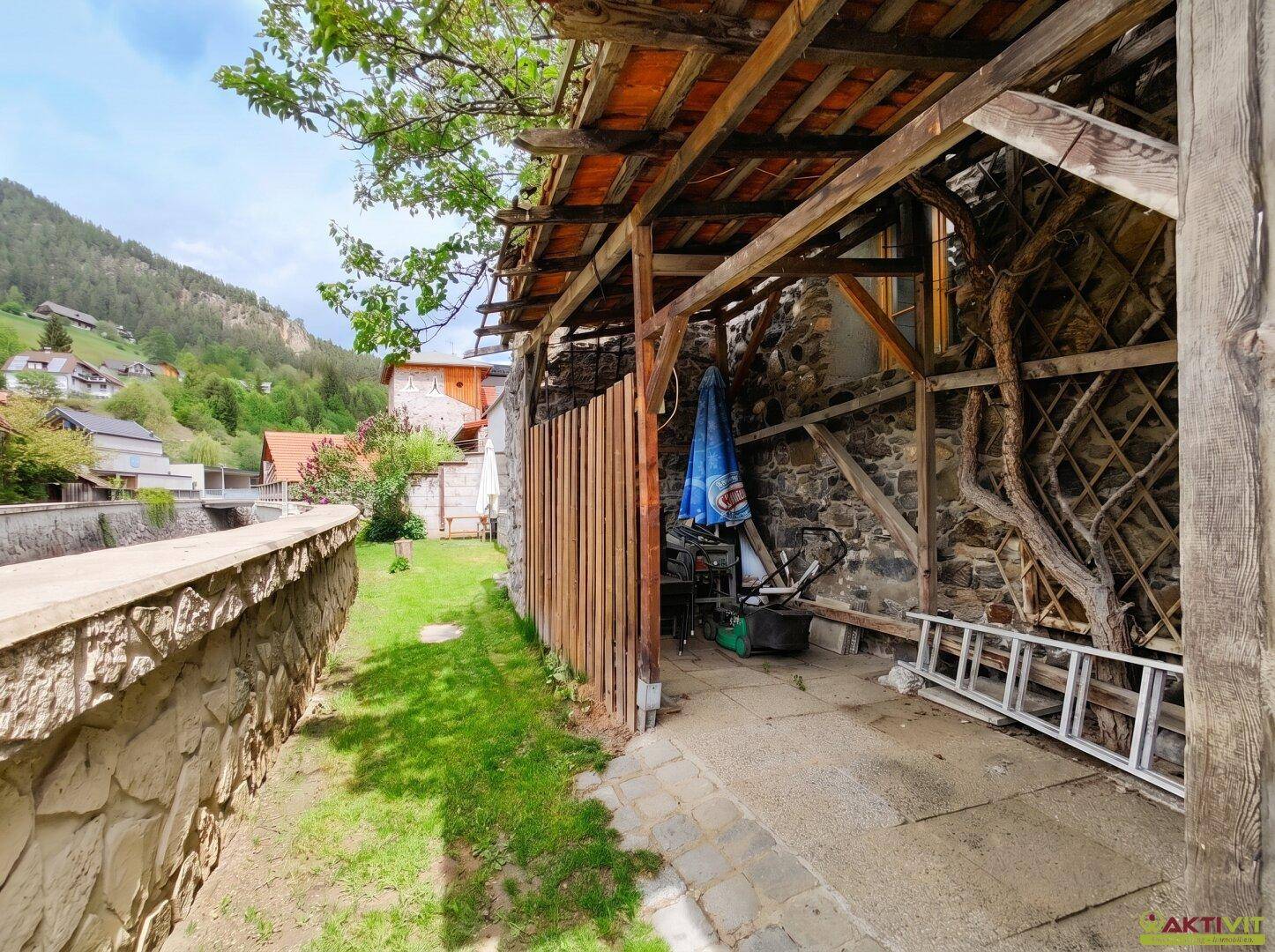 Seitlicher Gartenbereich mit überdachtem Unterstand, Steinmauer und viel Grün.