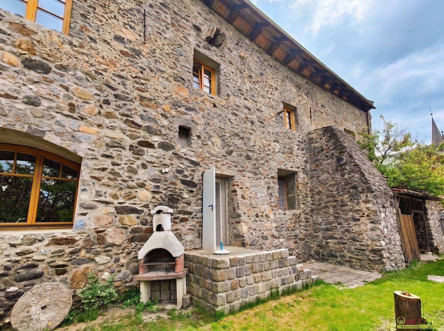 Steinfassade des Gebäudes mit Außengrill, grünem Rasen und Steintreppe.