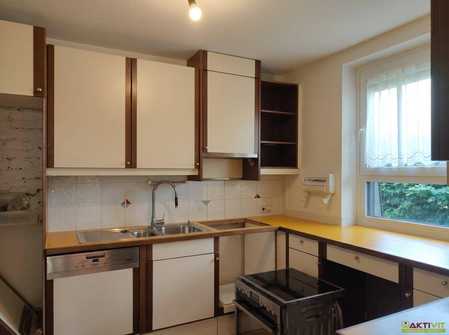 Ältere Einbauküche mit weißen Schränken, Spülmaschine und Fenster zum Garten.