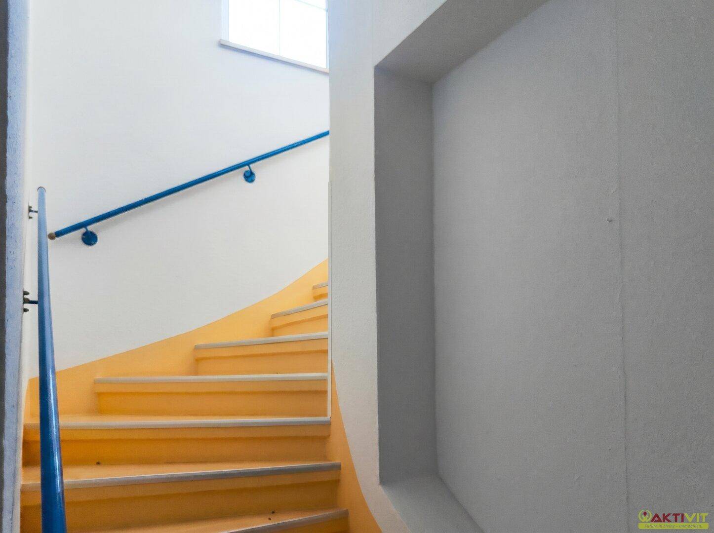 Treppenhaus mit gelb gestrichenen Stufen und blauem Handlauf, führt nach oben.