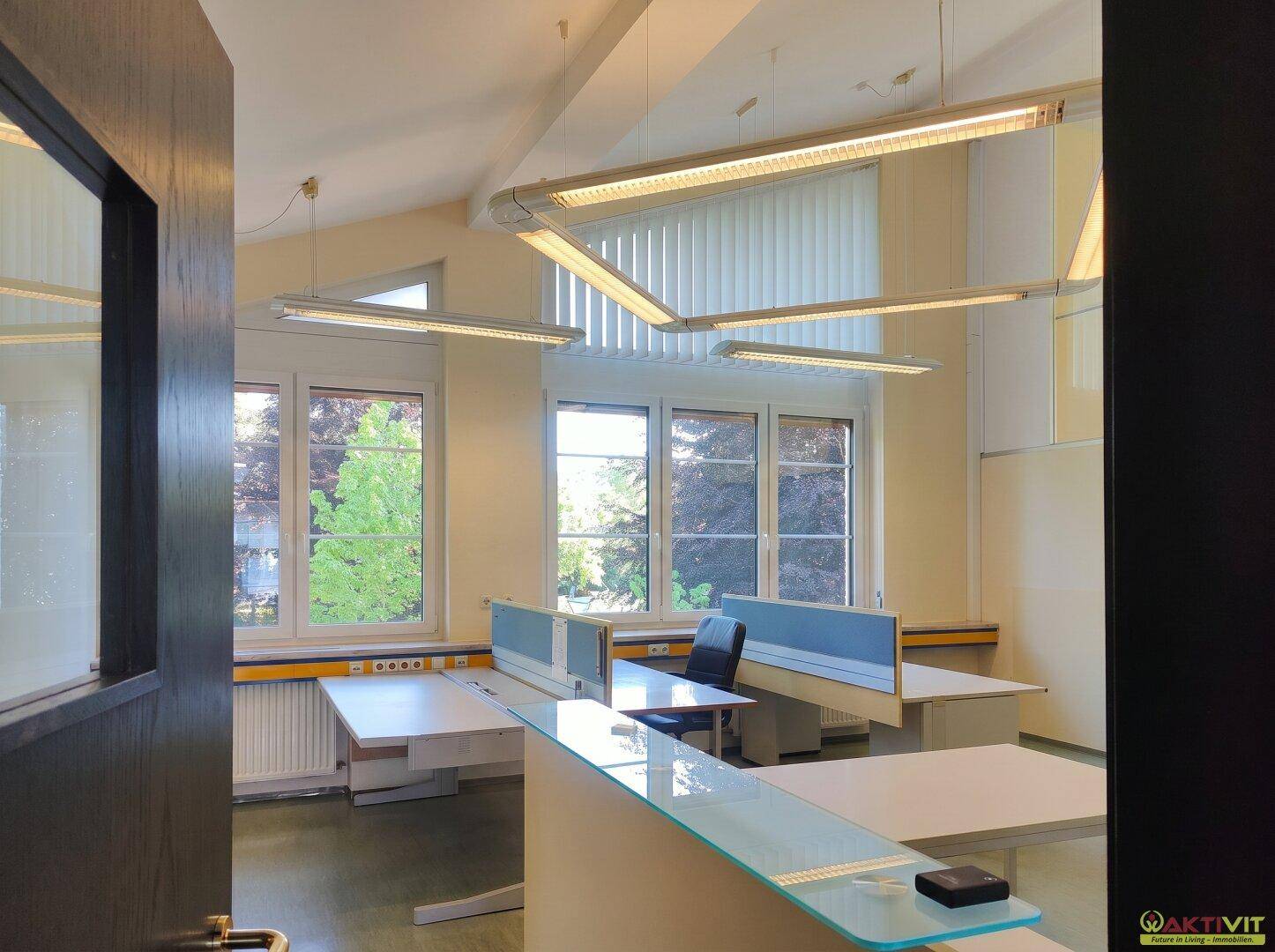 Helles Büro mit mehreren Arbeitsplätzen, großen Fenstern und modernen Deckenleuchten.