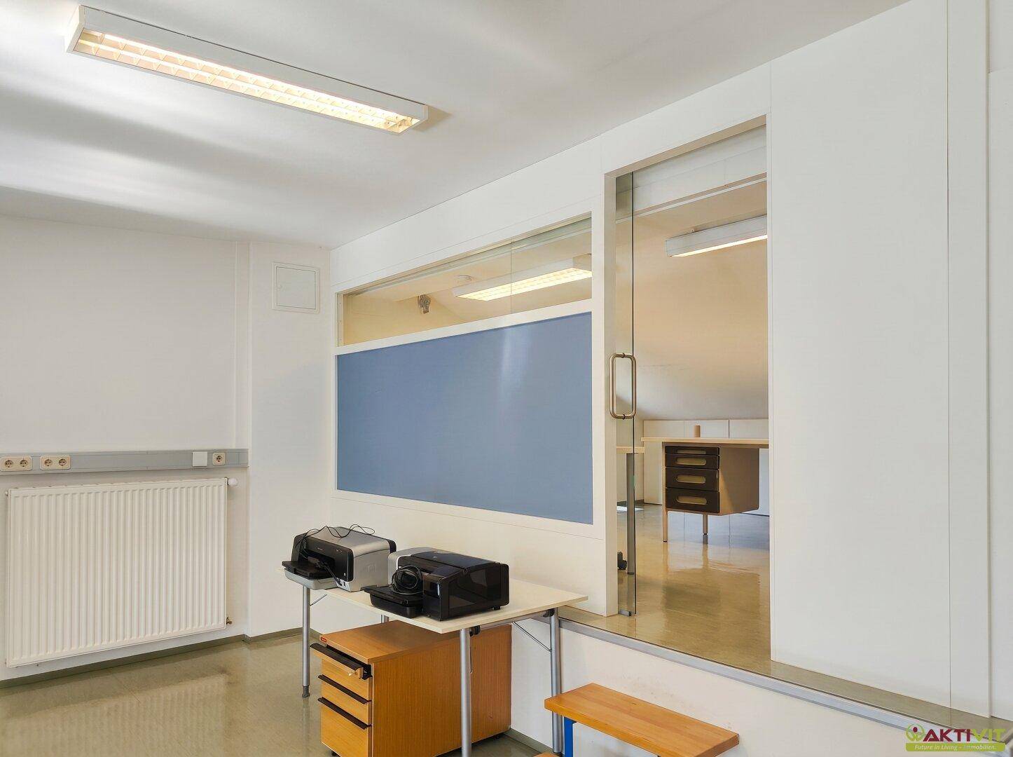 Bürobereich mit Druckerstation und Blick durch eine Glaswand in einen weiteren Raum.