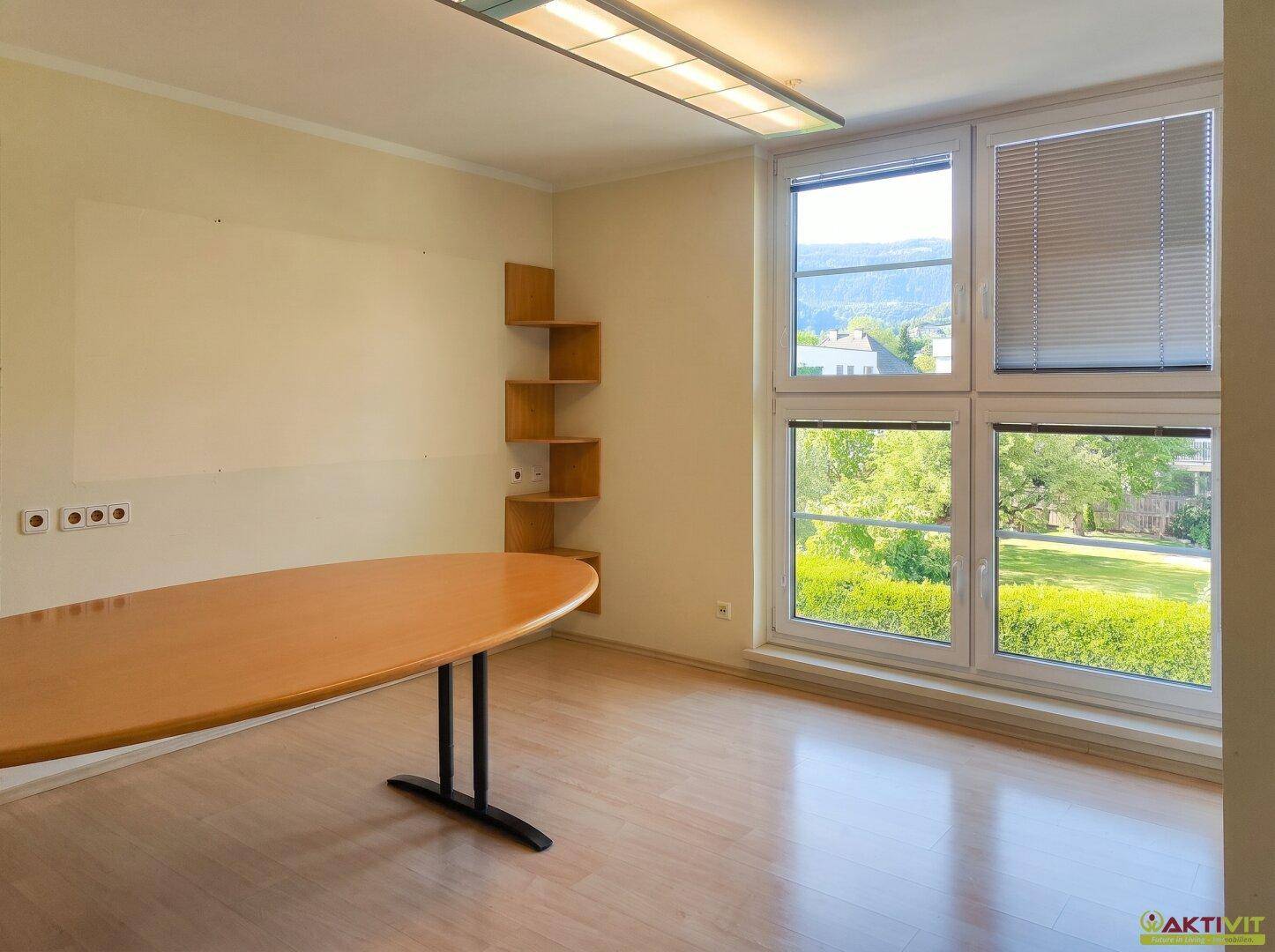 Büro mit Holztisch, Eckregal und großen Fenstern mit Blick ins Grüne.