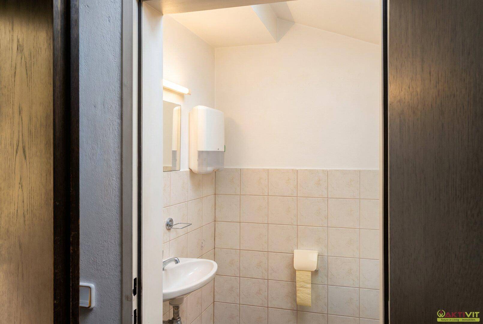 Kleine Toilette mit Waschbecken, hellen Fliesen und Spender für Papierhandtücher.