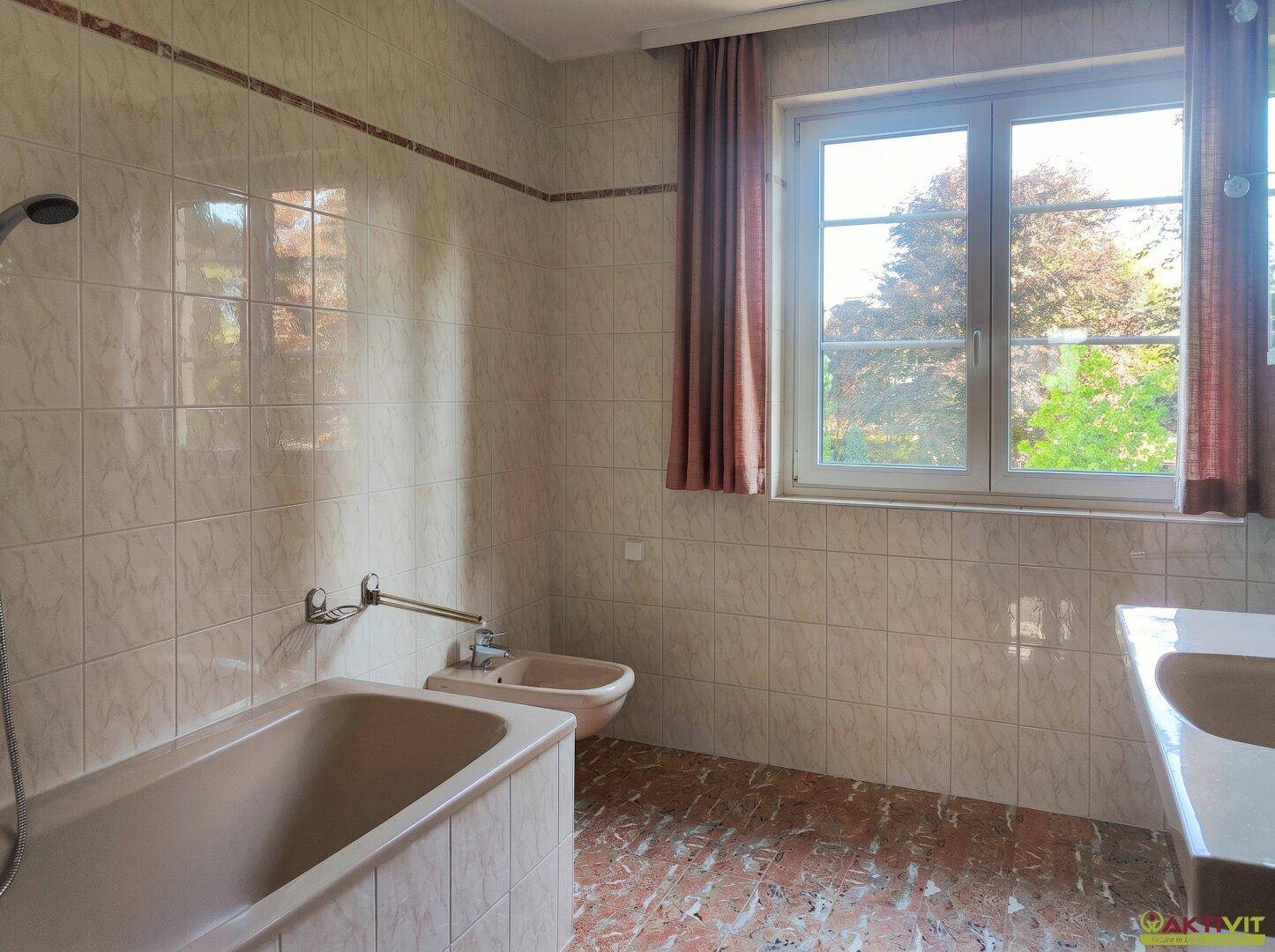 Badezimmer mit Badewanne, Bidet, Fliesenboden und Fenster mit Blick nach draußen.
